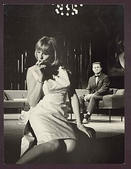 black and white photo of woman sucking her fingers while a man in a bowtie sits in the background. From a staging of Who's Afraid of Virginia Woolf by Edward Albee